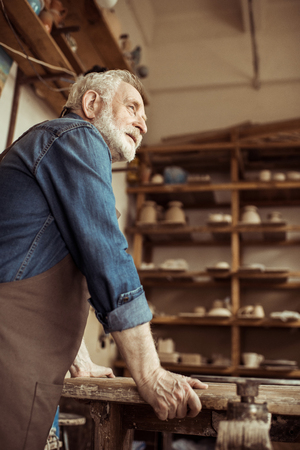 senior potter in apron standing and leaning on table against shelves with pottery goods at workshopの写真素材