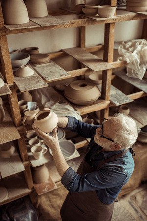 Senior potter in apron and eyeglasses examining ceramic bowlの写真素材