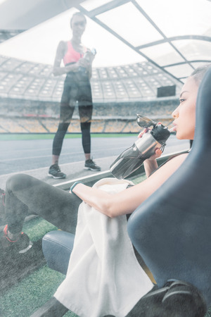 sportswoman with towel and bottle sitting on stadium seat and looking at sporty girlの写真素材