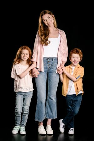 mother with adorable redhead children standing together and holding hands isolated on blackの写真素材