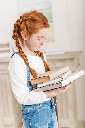 Portrait of adorable little redhead girl holding pile of booksの写真素材