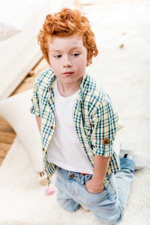 adorable little boy with hands in pockets kneeling on carpet and looking awayの写真素材