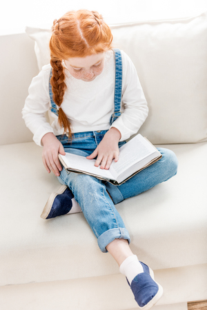 adorable little girl sitting on sofa and reading bookの写真素材