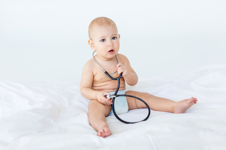 Adorable baby boy sitting on white bed with stethoscopeの写真素材
