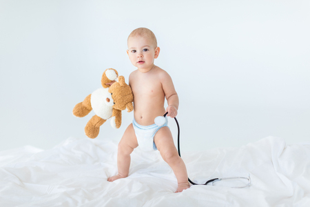Adorable small baby boy standing on bed and holding stethoscope with teddy bearの写真素材