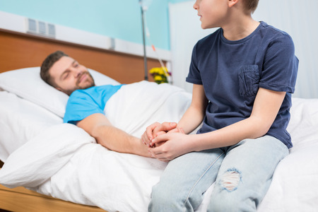 Cropped shot of son holding hand of sick father lying in hospital bedの写真素材