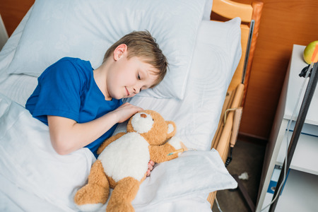 little boy with teddy bear lying in hospital bedの写真素材