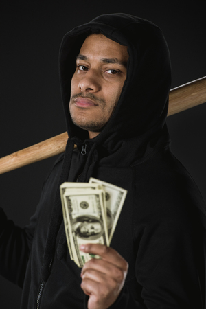 robber in zoodie with baseball bat and money isolated on blackの写真素材