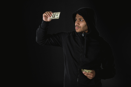 african american robber in zoodie holding money isolated on black