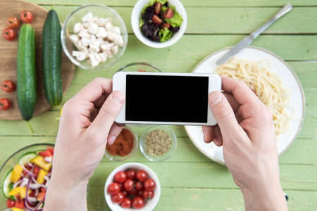 person holding smartphone with blank screen and photographing spaghetti and fresh vegetables on wooden tableの写真素材