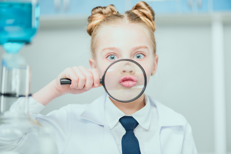 Little scientist in white coat holding magnifier in chemical lab