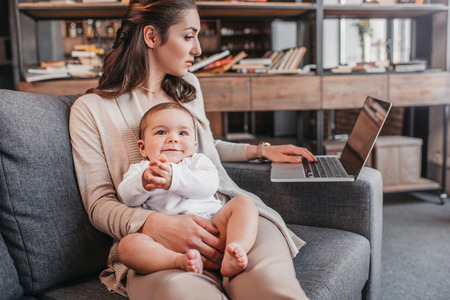 mother sitting on couch with her son and using laptop at homeの写真素材