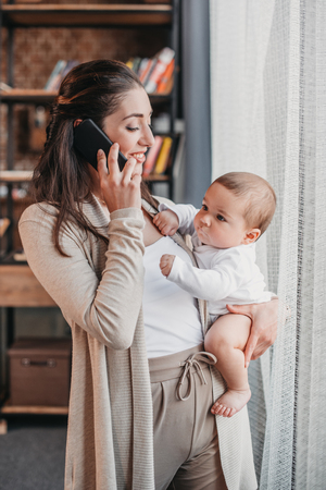 mother with little baby boy standing by the window and talking on smartphoneの写真素材