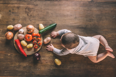 cute baby boy lying on kitchen table near group of vegetables on chopping boardの写真素材