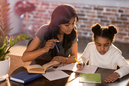 Mother and cute little daughter sitting at table and doing homework together at homeの写真素材