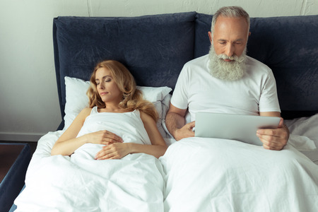 mature man using laptop in bed while blonde woman sleepingの写真素材