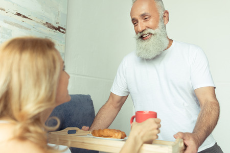 bearded man bringing tray with breakfast to happy blonde woman in bedroomの写真素材