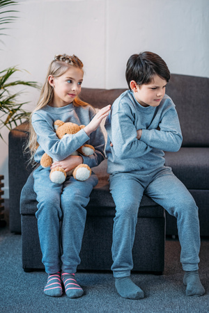 girl holding teddy bear while offended boy sitting on sofa at homeの写真素材