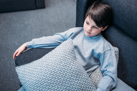 little boy in pajamas holding pillows while sitting on sofa and looking awayの写真素材