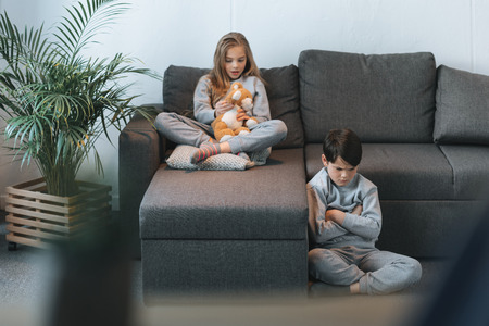 girl with teddy bear on sofa and boy sitting with arms crossedの写真素材
