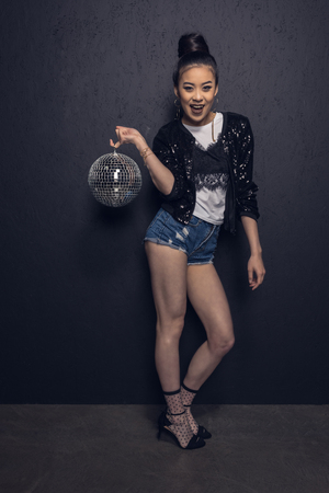 cheerful asian girl holding disco ball, looking at camera and posing at grey wall in studioの写真素材