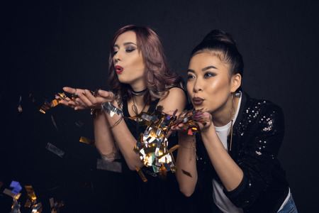 stylish young women blowing shiny confetti isolated on blackの写真素材