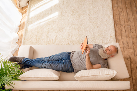 senior bearded man smiling and using digital tablet and lying on sofa at homeの写真素材