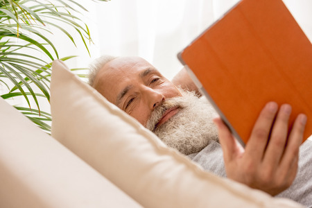 happy senior bearded man using digital tablet and lying on sofa at homeの写真素材