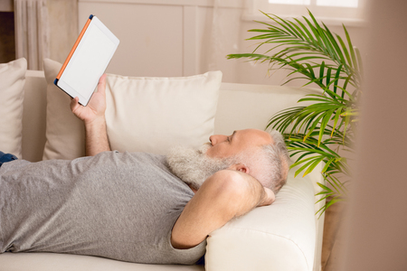 bearded man using digital tablet and lying on sofa at homeの写真素材