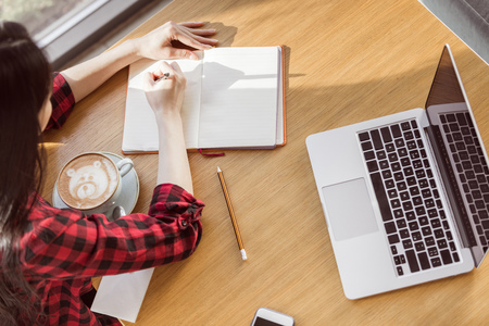 young woman sitting at table with coffee and laptop and writing in notebookの写真素材