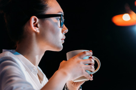 young businesswoman in eyeglasses holding cup while working late in officeの写真素材