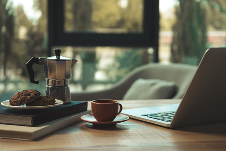 Close-up view of laptop, cup of coffee, moka pot and chocolate chip cookies with books on wooden tableの写真素材