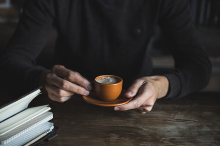 Close-up view of person drinking coffee while sitting at wooden table with notebook