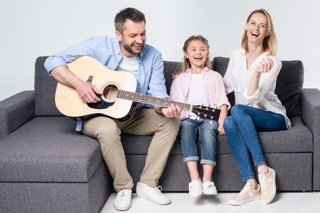 parents with cute little daughter sitting together on sofa and playing guitarの写真素材