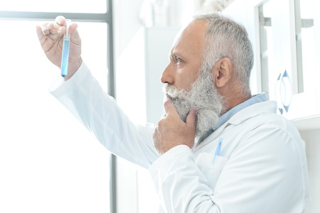 bearded scientist in white coat thinking and looking at tube with reagent in chemical lab