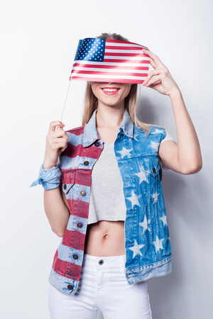 Smiling young woman in denim vest with stars and stripes hiding face by small american flagの写真素材