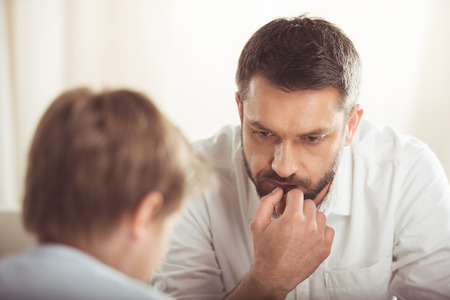 pensive bearded man with hand on chin looking away with son near byの写真素材