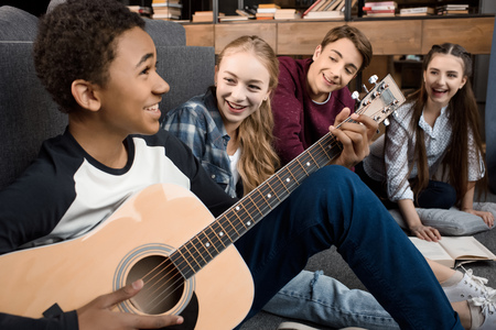 african american boy playing acustic guitar and singing while his friends listening at home