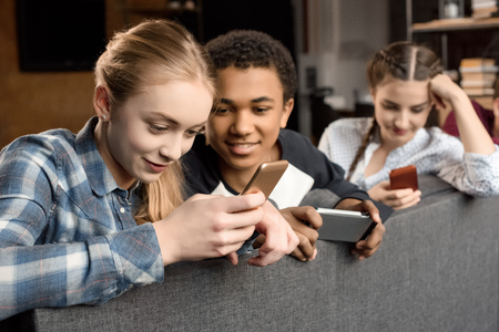 happy multicultural teenagers group using smartphones and sitting on sofa at home