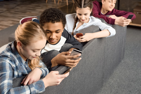 happy multicultural teenagers group using smartphones and sitting on sofa at homeの写真素材