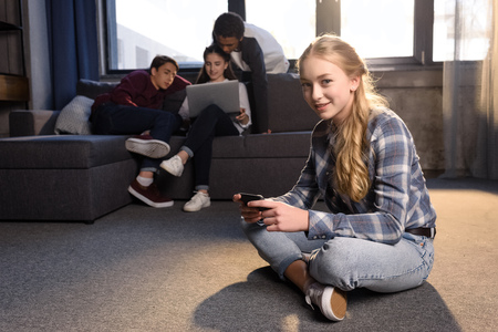 teenage girl sitting on floor and using smartphone with friends using laptop behindの写真素材