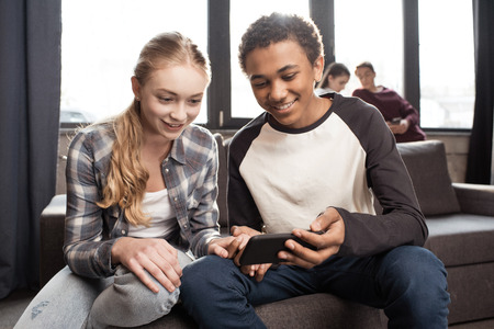 Happy teenage couple using smartphone on sofa while friends standing behindの写真素材