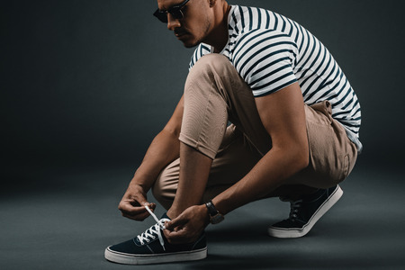 stylish middle aged man in striped t-shirt and sunglasses tying shoelaces in studioの写真素材