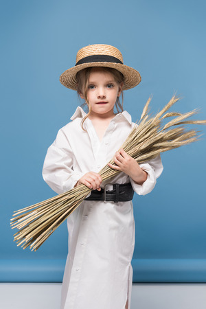 little girl in dress and straw boater holding wheat ears and looking at cameraの写真素材