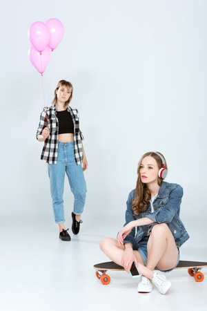 young woman in headphones holding smartphone and sitting on skateboard while pretty friend holding pink balloons behindの写真素材