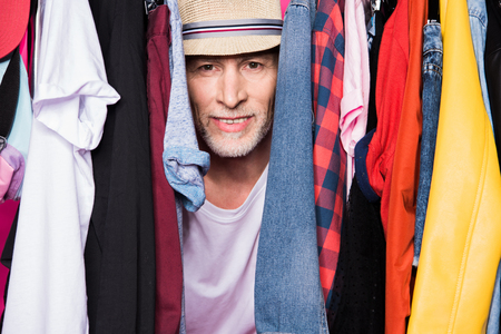 senior man wearing hat and standing with diferent clothes on hangers and looking at cameraの写真素材