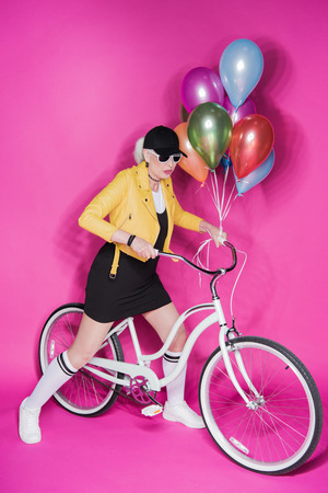 fashionable senior woman wearing yellow leather jacket holding colorful balloons and riding bicycleの写真素材