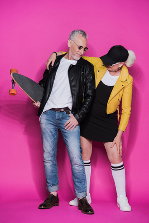 stylish senior couple posing together with skateboard isolated on pinkの写真素材