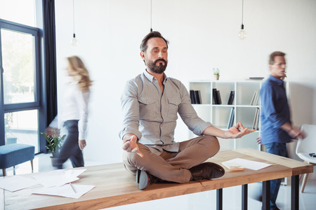 Relaxed businessman meditating in lotus position while coworkers movingの写真素材