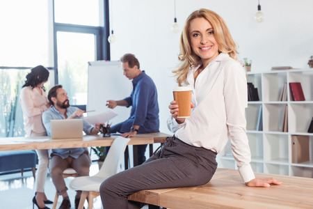 blonde businesswoman sitting on table and drinking coffee while colleagues working behindの写真素材